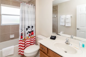 A bathroom with a Mickey Mouse themed shower curtain, a white toilet, a sink with a makeup towel, and a mirror reflecting towels.