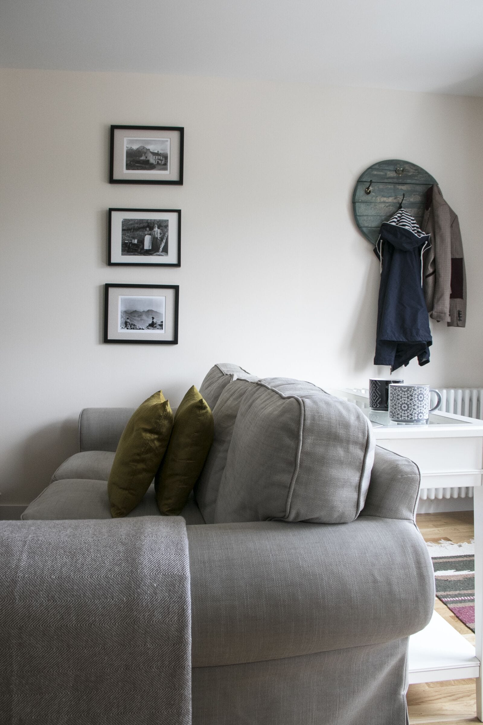 Un salon avec un canapé gris, trois tableaux encadrés accrochés au mur, un porte-manteau avec une veste, et une tasse à café posée sur une table d’appoint.