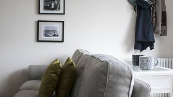 Un salon avec un canapé gris, trois tableaux encadrés accrochés au mur, un porte-manteau avec une veste, et une tasse à café posée sur une table d’appoint.
