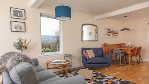 Una sala de estar con sofá, sillón, mesa de centro y una zona de comedor con mesa y sillas. Hay un ventanal con vistas, un espejo en la pared y cuadros enmarcados.