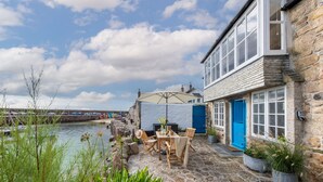 Een stenen gebouw met een blauwe deur, een terras met een tafel en stoelen, en uitzicht op het water.