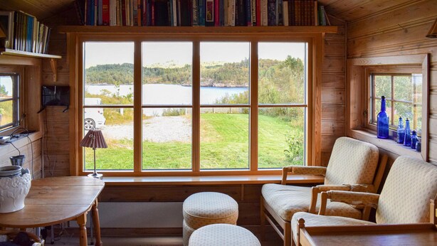 L’intérieur d’un chalet accueillant, doté d’une grande fenêtre offrant une vue sur un lac et une forêt. On y trouve des étagères en bois, une table avec un vase et deux coins salon confortables.
