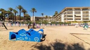 Ein Strand mit einer blauen Spielkonstruktion, Palmen und einem mehrstöckigen Hotel im Hintergrund.