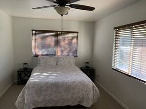 Un dormitorio con cama, dos mesitas de noche, un ventilador de techo y dos ventanas con persianas.