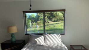 Un dormitorio con cama, una mesita de noche con lámpara, una ventana con persianas y una pequeña mesa.