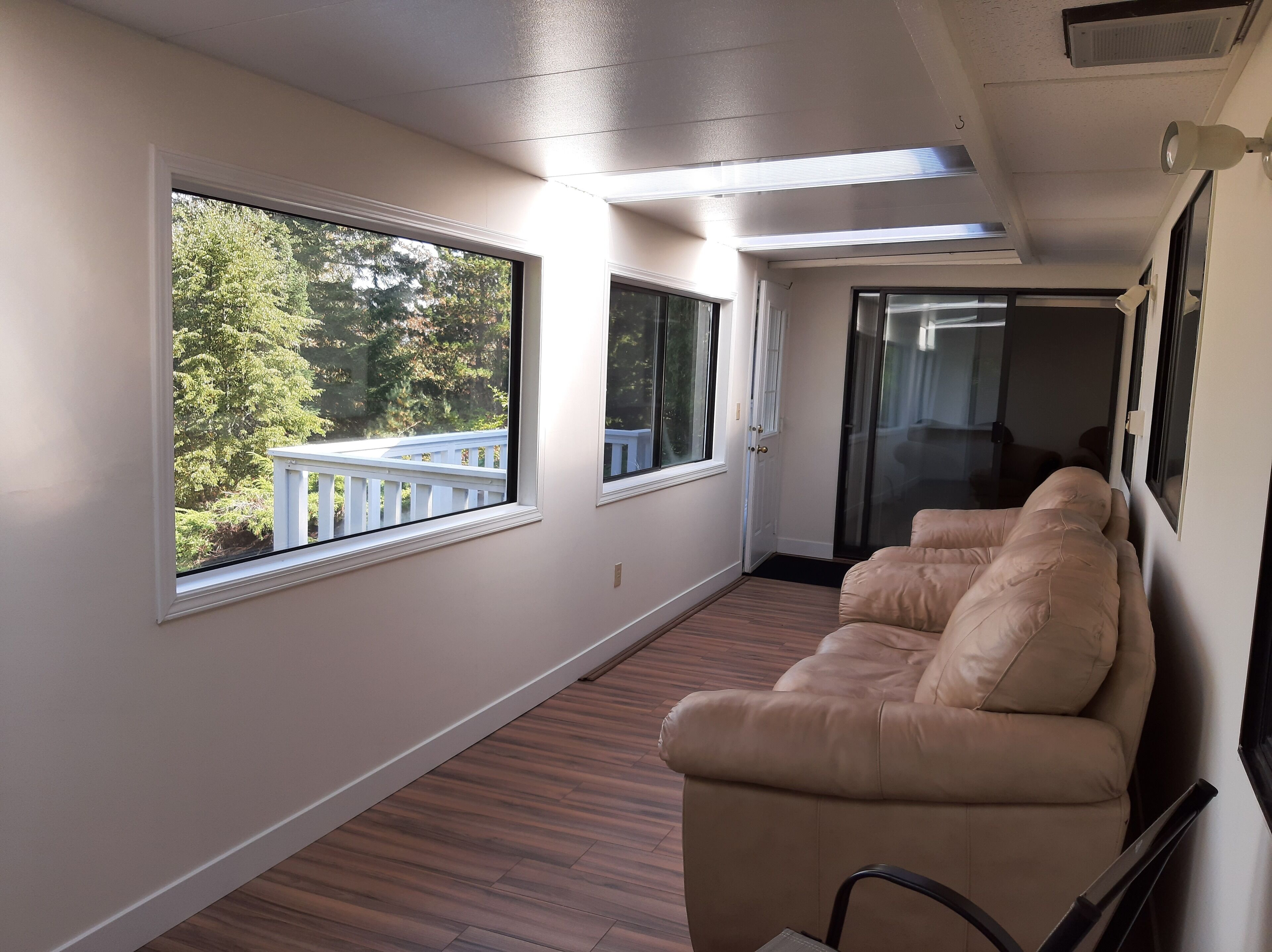 A corridor with a beige sofa, wooden flooring, and large windows offering a view of a balcony and trees.