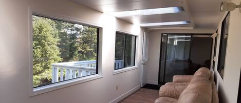 A corridor with a beige sofa, wooden flooring, and large windows offering a view of a balcony and trees.