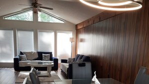 Un salon moderne avec une table à manger en verre, des fauteuils en cuir, un ventilateur de plafond et de grandes fenêtres équipées de stores.