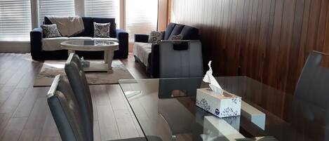 A modern living room with a glass dining table, leather chairs, a ceiling fan, and large windows with blinds.
