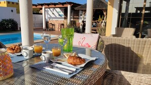 A breakfast setup with croissants, orange juice, and coffee on a wicker table by a poolside. - 4 bedroom LUXURY villa with AC, private heated pool, XBOX and all the amenities! (Castillo Caleta de Fuste)
