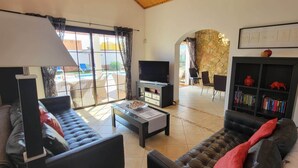 A living room with a sofa, a coffee table, a TV, and a bookshelf. There is a large window with curtains, a stone wall, and a view of the outside. - 4 bedroom LUXURY villa with AC, private heated pool, XBOX and all the amenities! (Castillo Caleta de Fuste)