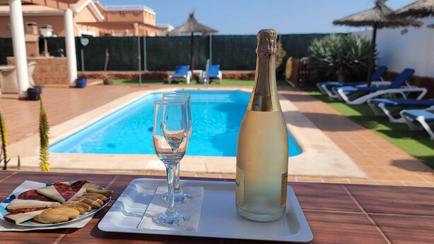 A table with a bottle of champagne, a glass, and a plate of snacks by a poolside. - 4 bedroom LUXURY villa with AC, private heated pool, XBOX and all the amenities! (Castillo Caleta de Fuste)