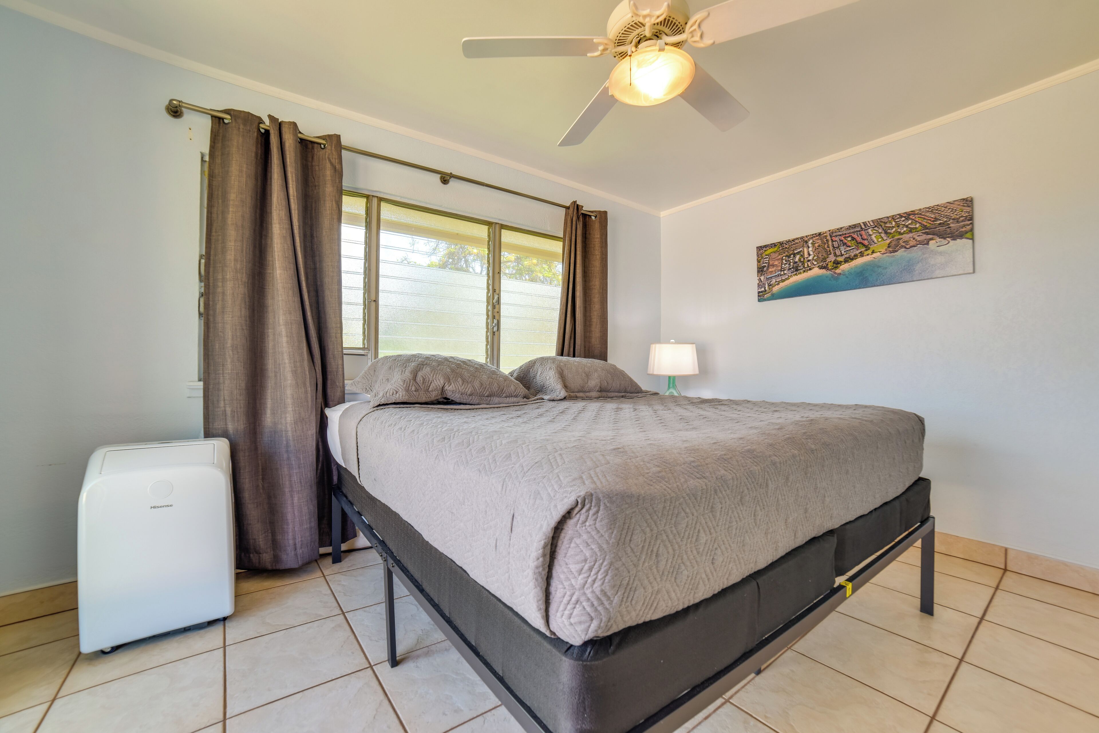 A bedroom with a bed, a ceiling fan, a window with curtains, a small white appliance, and a wall-mounted lamp.