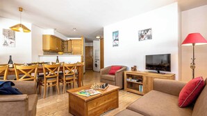 Ein modernes Wohnzimmer mit Couch, Fernseher und Couchtisch. Ein Essbereich mit Holztischen und -stühlen. Der Raum zeichnet sich durch ein klares, minimalistisches Design mit weißen Wänden und Holzböden aus.