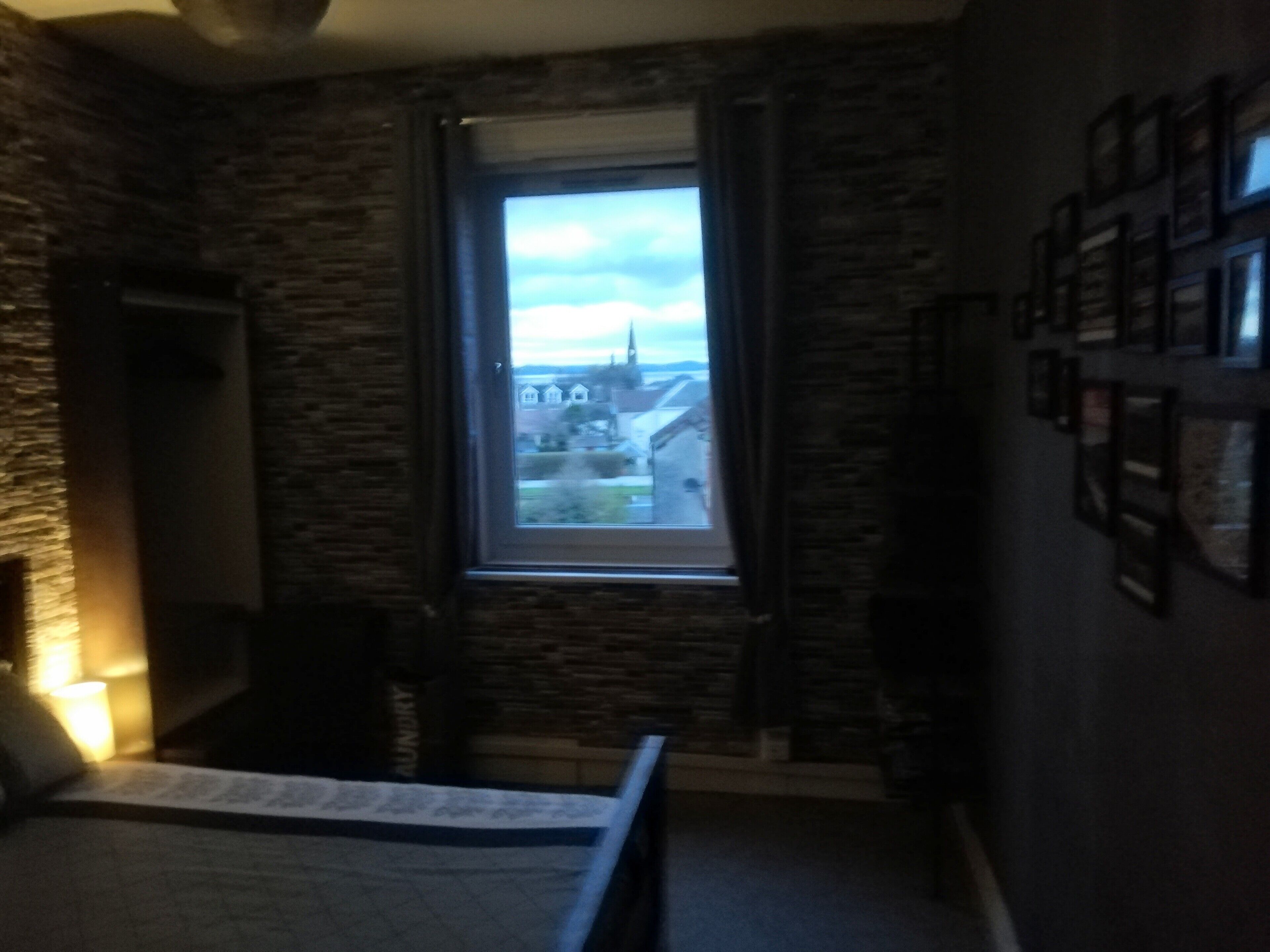 A bedroom with a stone wall, a window with a view of houses, and a bed.