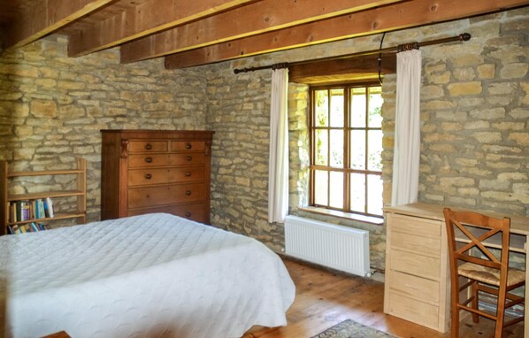 Un dormitorio con pared de piedra, vigas de madera en el techo, cama, cómoda, silla y ventana con cortinas.