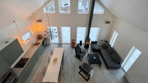 A spacious loft with a high ceiling, a central fireplace, a dining area, and large windows offering views of a snowy landscape.
