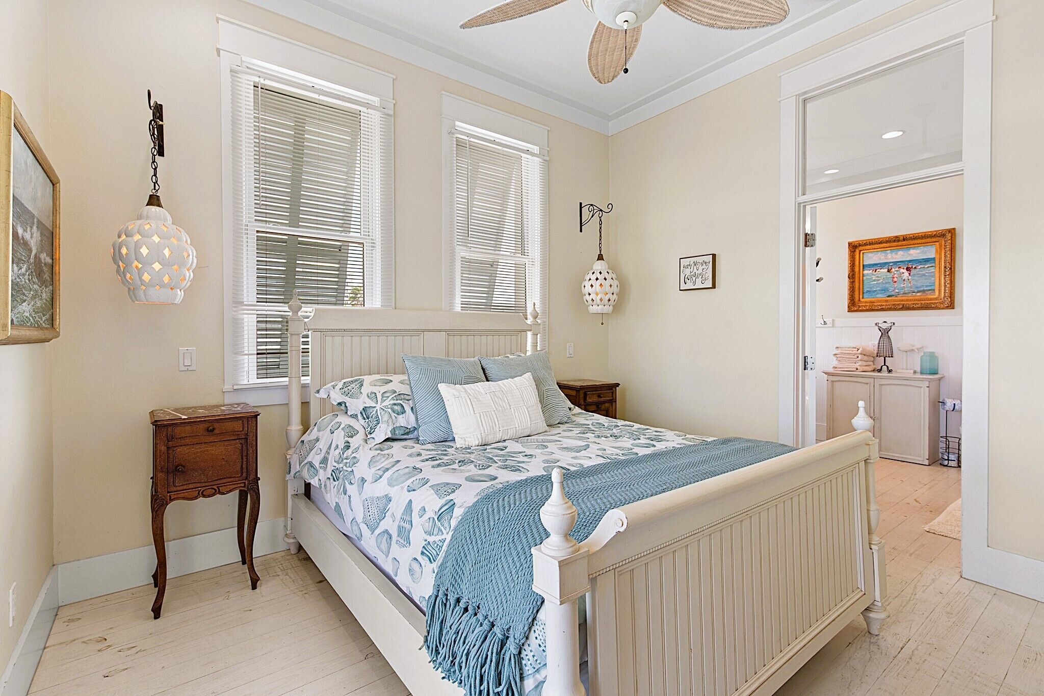 A bedroom with a bed, two windows with blinds, a bedside table, a lamp, a wall clock, and a door leading to another room.