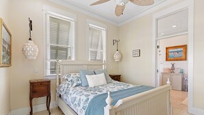 A bedroom with a bed, two windows with blinds, a bedside table, a lamp, a wall clock, and a door leading to another room. - Lotus Villa-Beautiful Straw paths through the pretty dunes. Look out tower! (South Padre Island)
