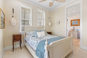 Un dormitorio con cama, dos ventanas con persianas, una mesita de noche, una lámpara, un reloj de pared y una puerta que da a otra habitación.