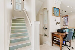 A staircase with a white railing, a desk with a chair, a painting on the wall, and a window with blinds.