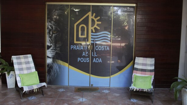 A hotel entrance with a large glass door featuring a logo and text. There are two lounge chairs with cushions outside. - Pousada praia de costazul (Rio das Ostras)
