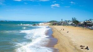 A beach with a sandy shore, waves, and buildings in the background. - Pousada praia de costazul (Rio das Ostras)