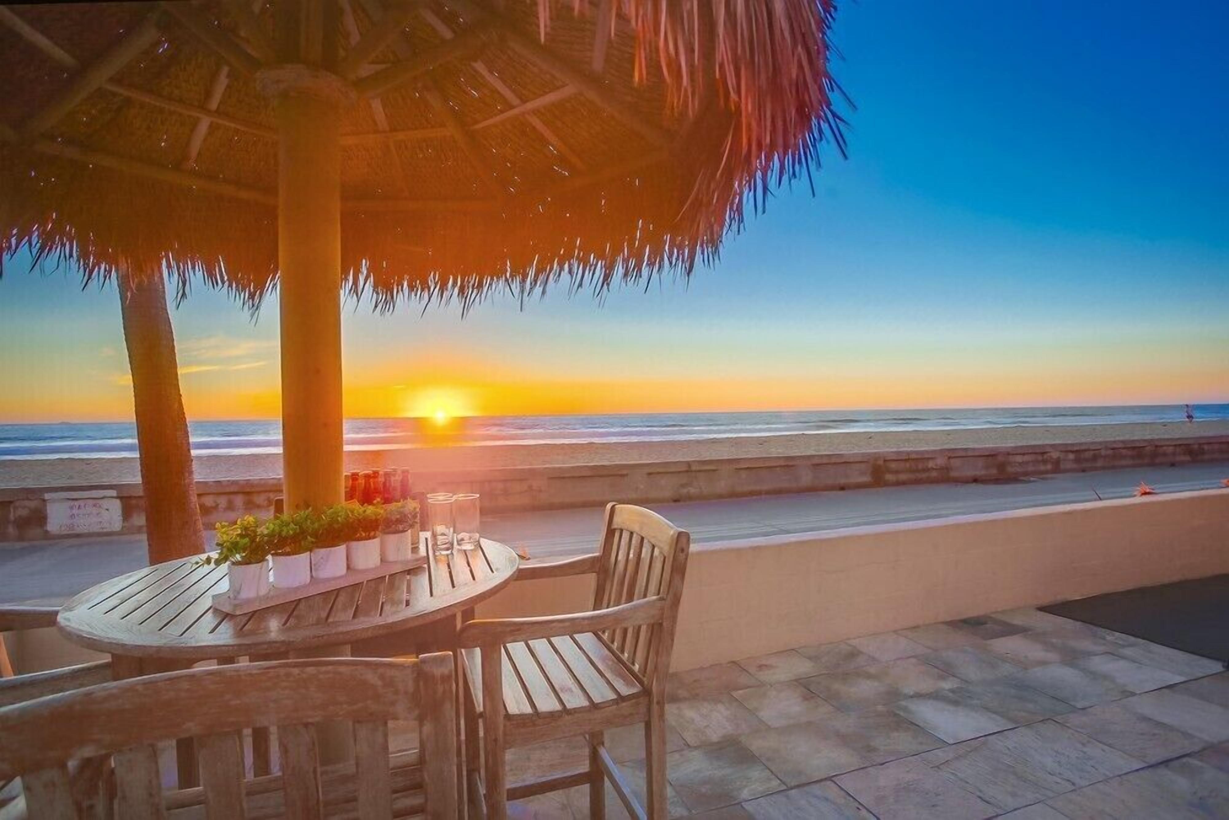 Un espace extérieur aménagé avec une table et des chaises en bois, un parasol en chaume et une vue sur le coucher de soleil sur l’océan.