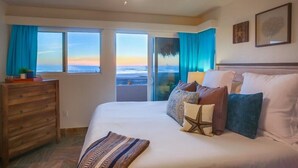 Una camera da letto con un letto grande, un ventilatore a soffitto e vista sull'oceano attraverso ampie finestre.