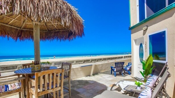 Una veranda fronte spiaggia con ombrellone di paglia, poltrone relax e un tavolo con sedie.