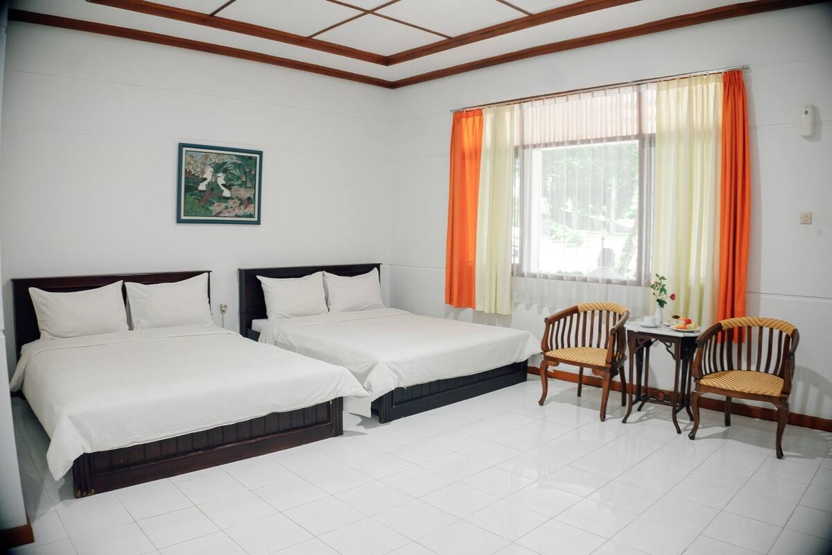 Een hotelkamer met twee eenpersoonsbedden, een kleine tafel met stoelen, een raam met oranje gordijnen en een schilderij aan de muur.