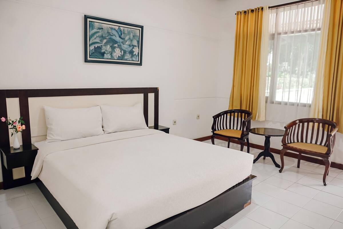 Een hotelkamer met een bed, twee stoelen, een klein tafeltje, een schilderij aan de muur en een raam met gordijnen.