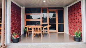 A covered outdoor seating area with a table and chairs, surrounded by red lattice panels and potted plants.
