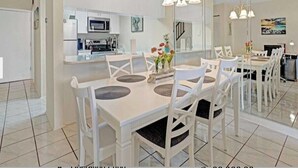A modern dining area with a white table and chairs, a chandelier, and a kitchen in the background. - Turtle Oasis - 2Bd 2B Townhouse (ST. AUGUSTINE)