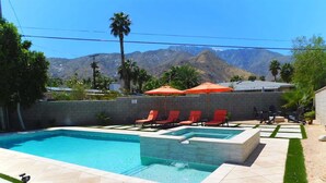 A swimming pool with red lounge chairs and an umbrella, surrounded by a stone wall and a view of mountains. - Experience Modern Luxury at Desert Hideaway (Palm Springs)