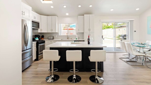 A modern kitchen with white cabinets, stainless steel appliances, and a central island with bar stools. - Experience Modern Luxury at Desert Hideaway (Palm Springs)