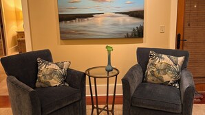 Una sala de estar con dos sillones grises, una mesita redonda con un florero, y un cuadro grande de paisaje en la pared.