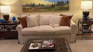 Una sala de estar con un sofá color beige, dos mesitas de noche con lámparas, una mesa de centro con revistas y un cuadro de paisaje grande en la pared.