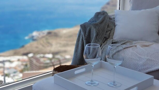 A room with a view of the sea, a bed with a white pillow, a grey blanket, and two glasses on a tray.