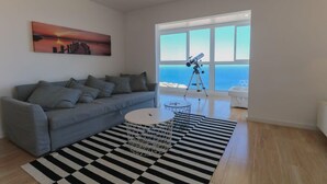 A modern living room with a grey sofa, a black and white striped rug, a telescope, and a large window with a view of the sea.