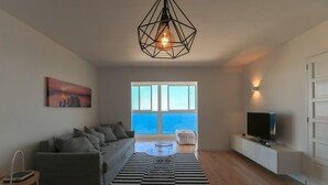 A modern living room with a sofa, a coffee table, a TV, and a large window with a view of the sea.