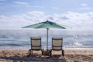 Une plage avec deux chaises sous un parasol vert, un panier, et l’océan en arrière-plan.