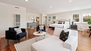 Una habitación moderna y bien iluminada con una cama, un sofá, una silla y una mesita con una botella y un vaso.