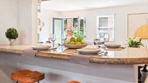 Una cocina moderna con encimeras de granito, una isla central con un cuenco de fruta y vistas al exterior a través de un ventanal.