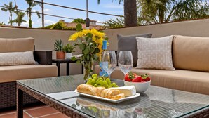 Una mesa de vidrio con un plato de queso, uvas y fresas, una botella de vino y un jarrón con girasoles en un patio exterior con un sofá y una pequeña mesa.
