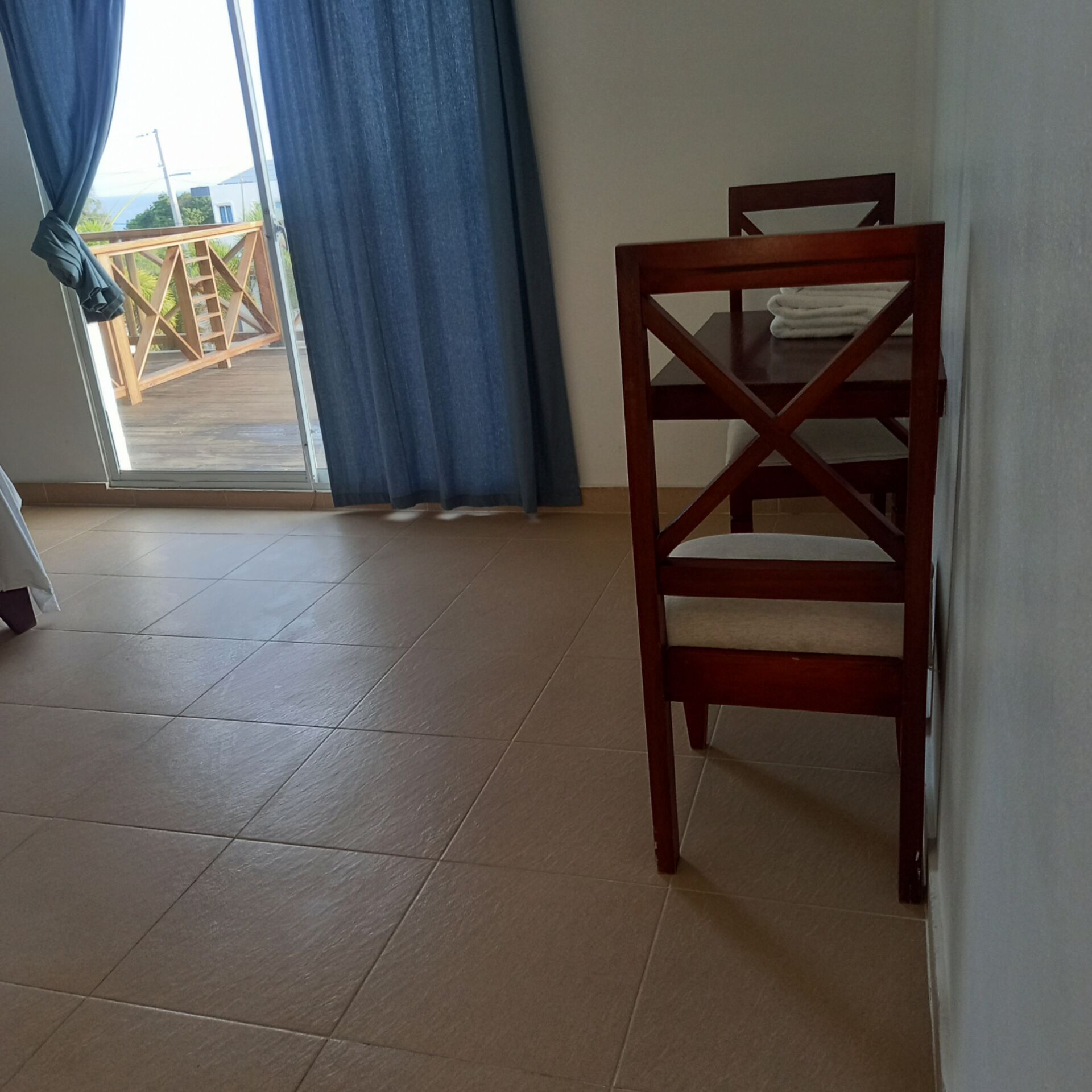 A room with a wooden dining table and chairs, a tiled floor, and a view through a glass door with blue curtains.