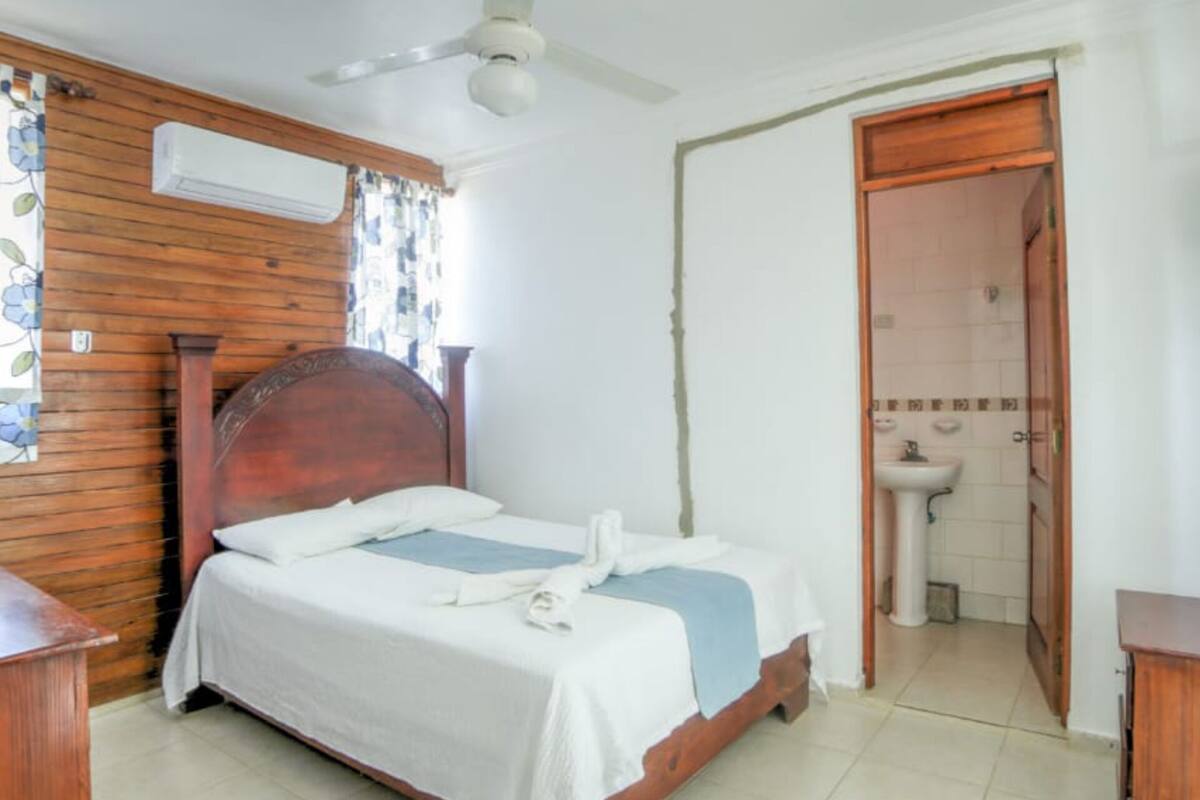A bedroom with a wooden bed, a ceiling fan, a bathroom with a sink and mirror, and a door leading to another room.