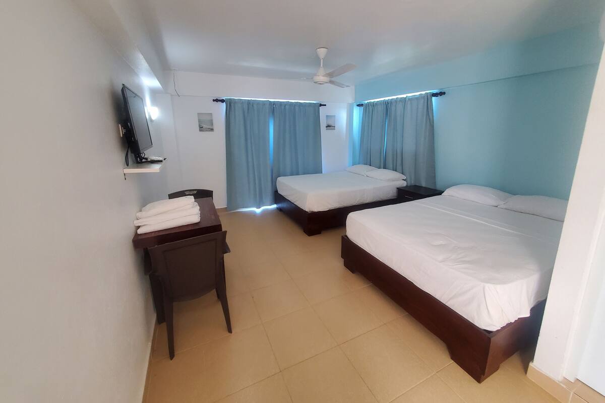 A hotel room with two beds, a desk, a chair, a TV, and a ceiling fan.