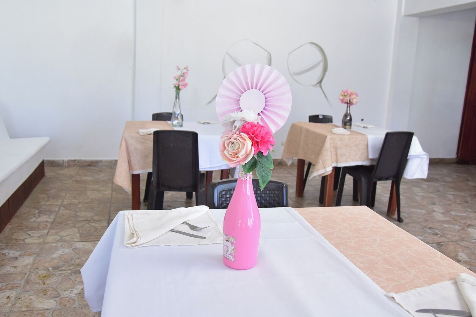 Een eetgedeelte met tafels voor twee personen, elk met een wit tafelkleed, een roze vaas met bloemen en een ventilator.
