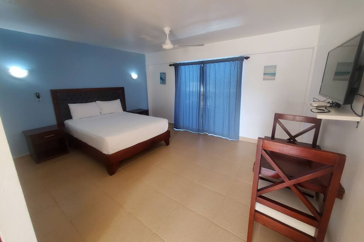 A bedroom with a bed, a nightstand, a desk with a chair, a ceiling fan, and a window with curtains.